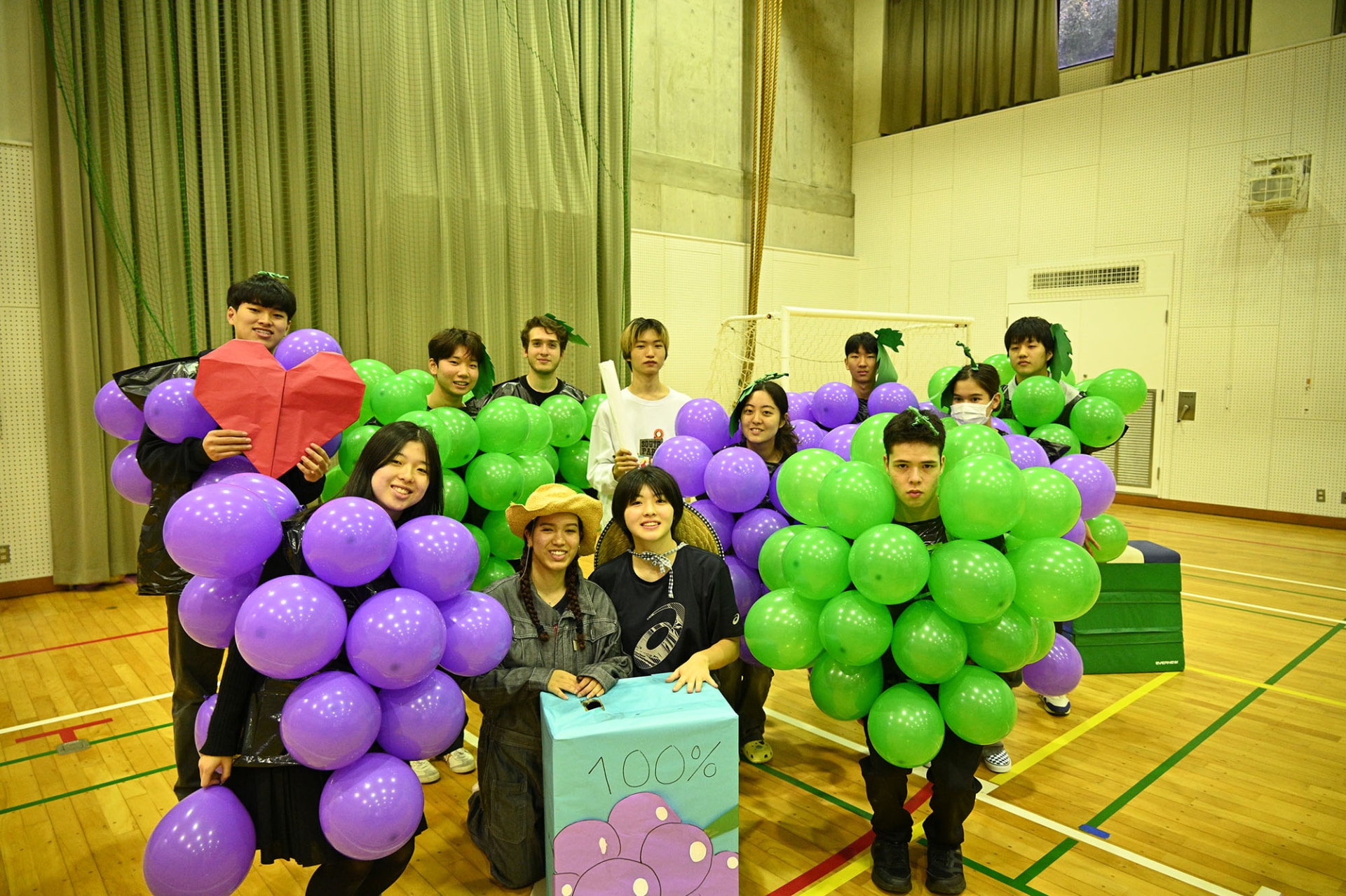 Students and adults holding clusters of purple and green balloons in a gymnasium, posing for a group photo.