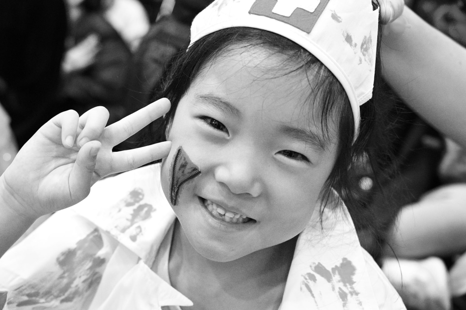 Smiling child wearing a paper hat and face paint, holding up a peace sign.