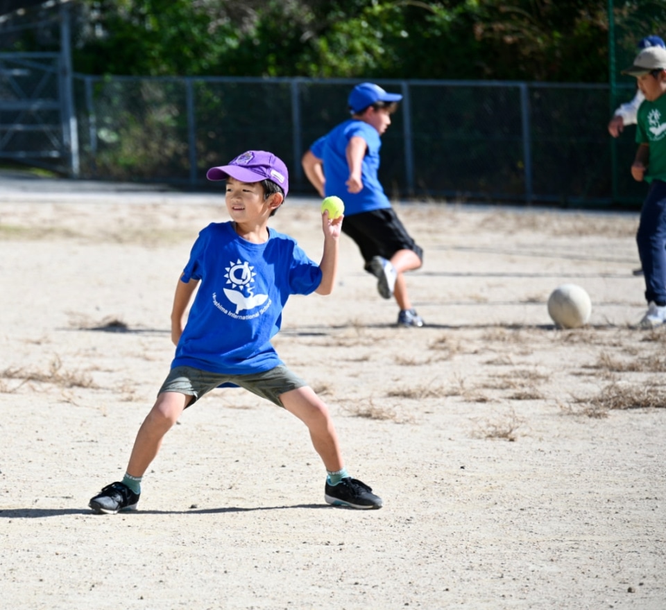 Young child in a blue shirt and purple cap fielding a ball on a sandy playground during a game.