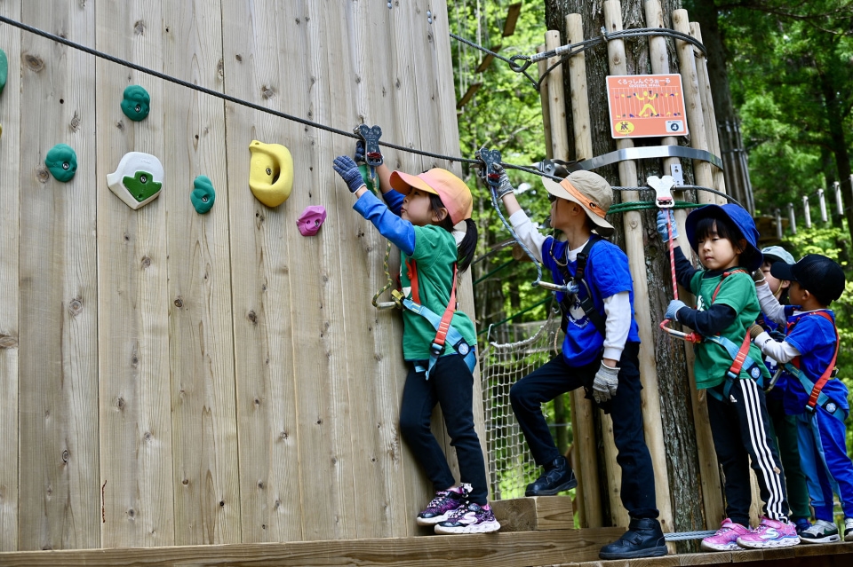 Children wearing helmets and harnesses climbing an outdoor rock wall at an adventure course.