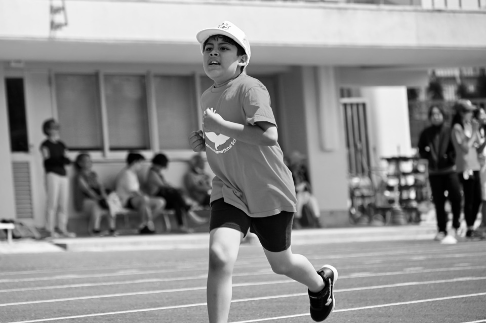 Young boy running on an outdoor track during a school race with spectators in the background.
