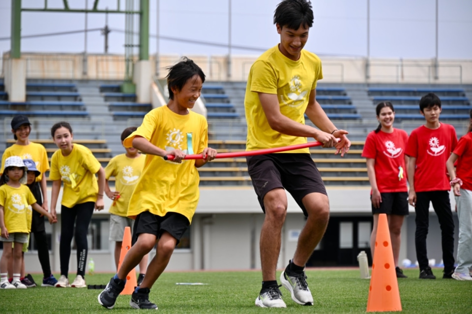 Adult and child running together during a relay race on a sports field with teammates cheering nearby.