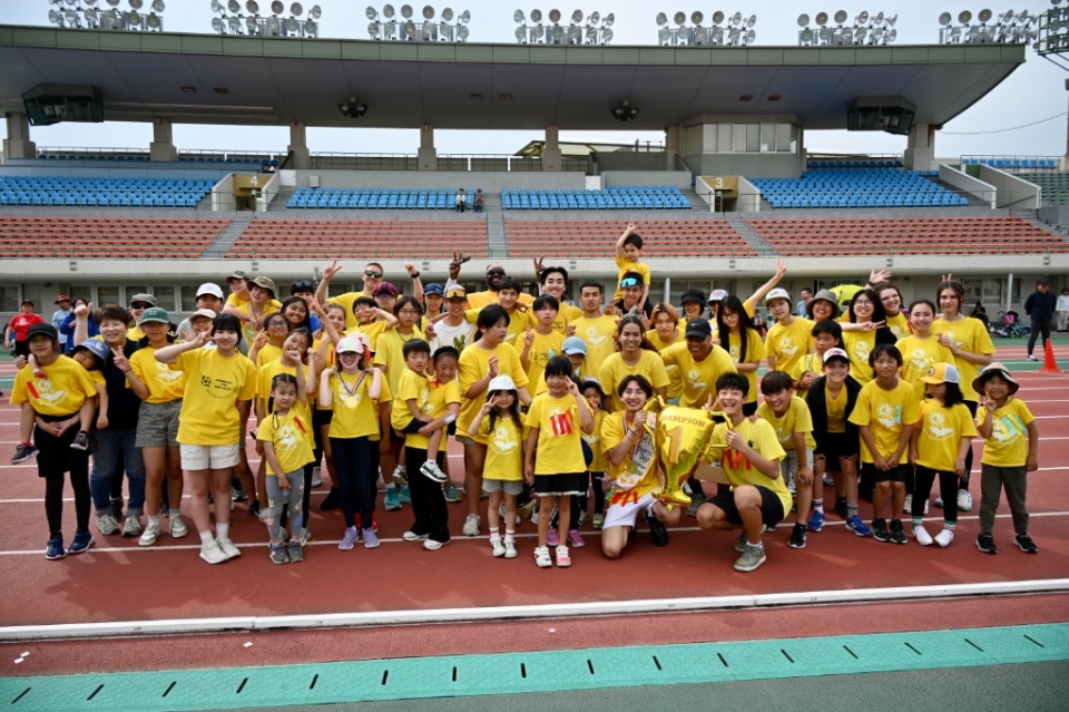 Large group of students and adults in yellow shirts posing together on a stadium track during a school event.